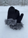 A pair of black knitted mittens with gray faux fur trim on the cuffs, displayed on a snowy surface.