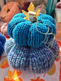 A two-tier, multi-colored blue knit stuffed pumpkin with a wooden stem and decorative fabric leaves on a table with a variety of other textiles.