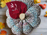 Knitted pumpkin decoration with a red reindeer face on a wooden surface with autumn leaves.