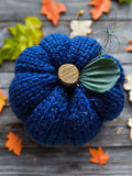 Knitted blue pumpkin decoration with a wooden stem on a wooden surface with autumn leaves.