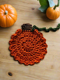 A crocheted orange pumpkin-shaped coaster with a green stem on a beige tabletop with pumkins