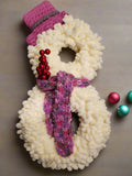 A decorative yarn snowman wreath featuring a cream-colored snowman with a long pink scarf and pink hat, with a white base covered in multi-colored yarn.